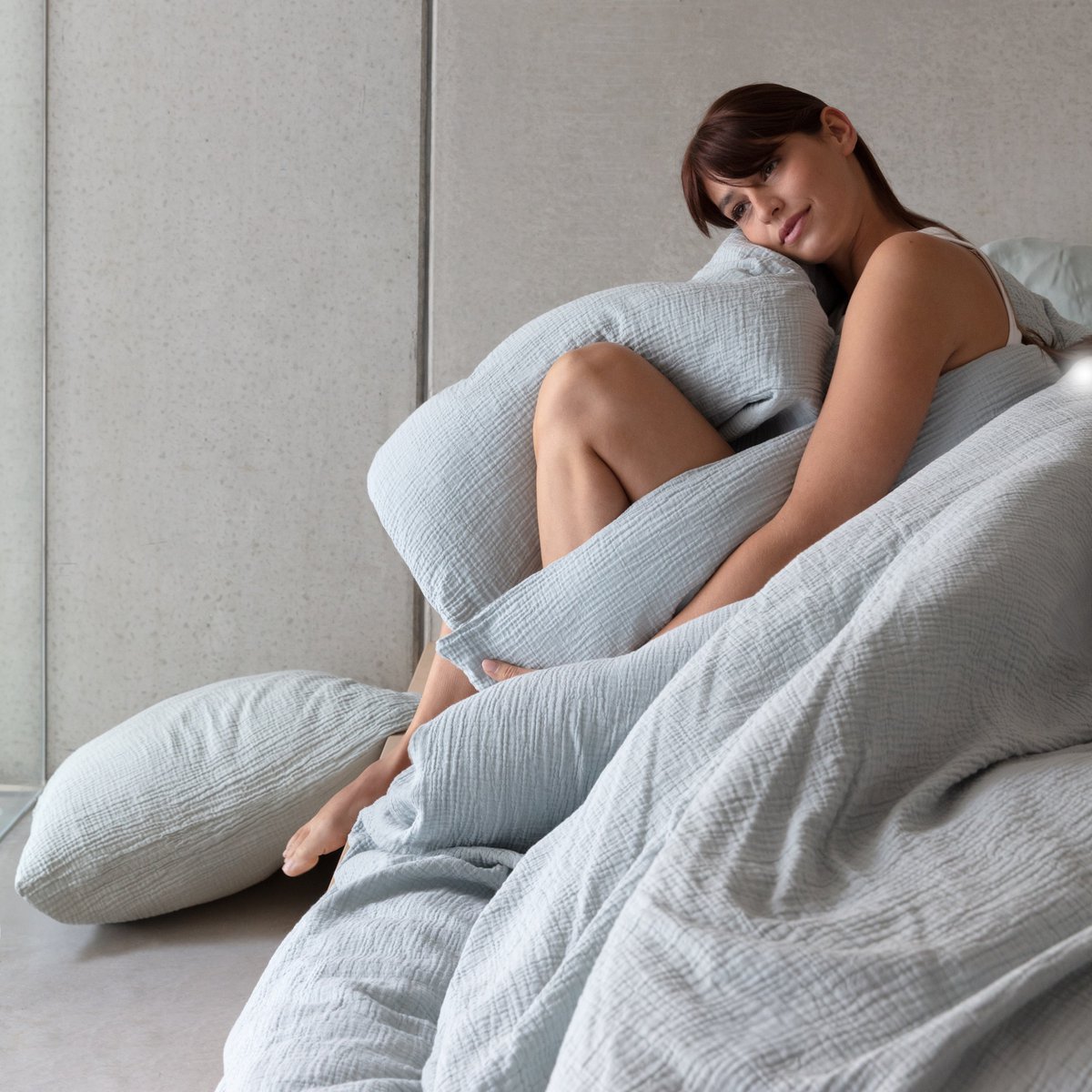 Woman sitting against misty blue cotton muslin pillows and a duvet cover with visible wrinkled texture