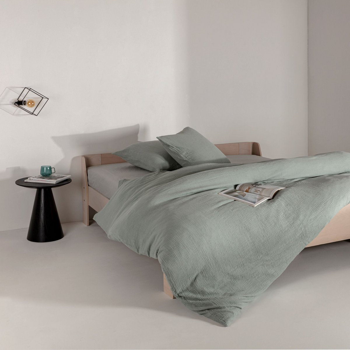 Sage green cotton muslin duvet cover and pillowcases styled on a bed with a magazine