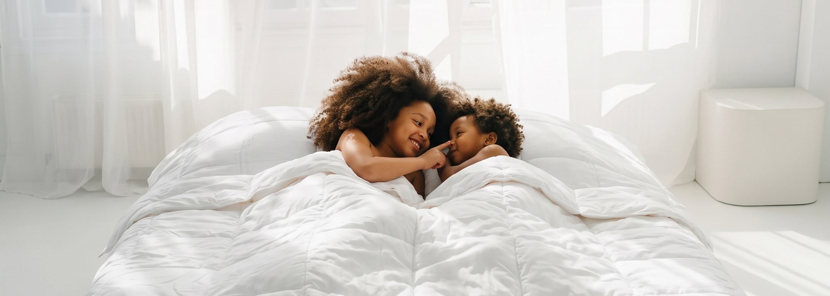 Two joyful children cuddling beneath a cozy white blanket on a bed bathed in natural sunlight, showcasing premium home textiles.