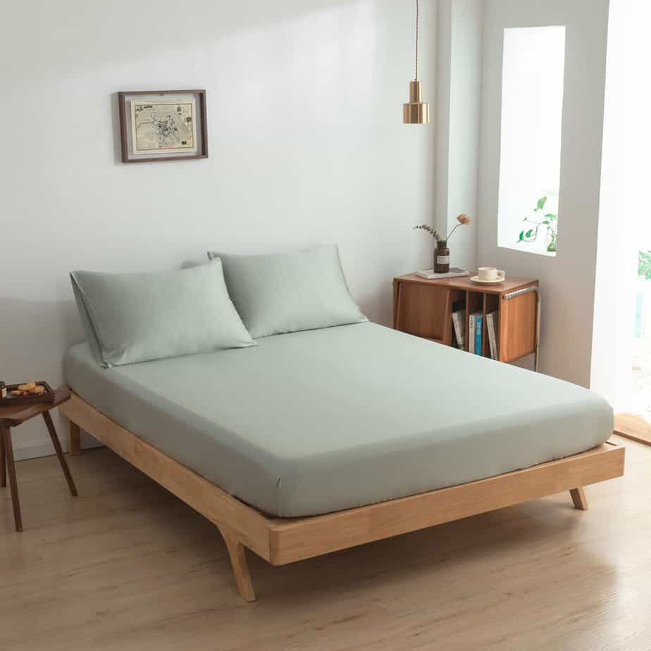 Sage Green bamboo fitted sheet styled on a wood-frame bed by a window, with matching pillowcases.
